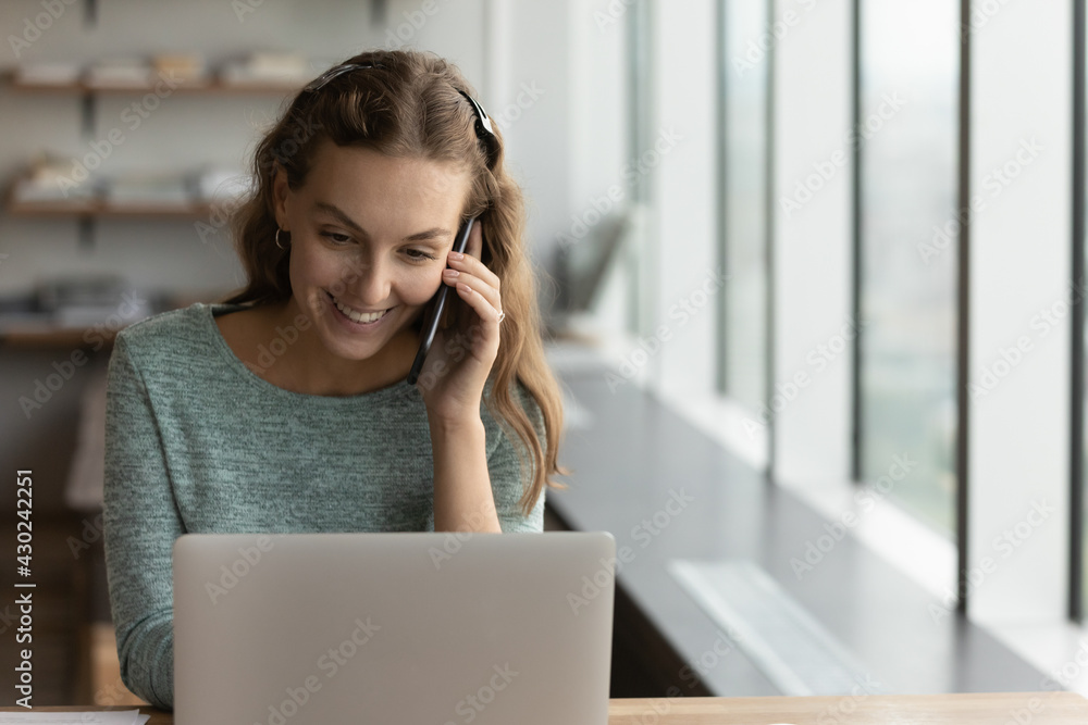 Happy young Caucasian businesswoman work on laptop in office talk speak on cellphone call with business partner. Smiling female employee worker have online consultation on smartphone using laptop. Happy young Caucasian businesswoman work on laptop in office talk speak on cellphone call with business partner. Smiling female employee worker have online consultation on smartphone using laptop.