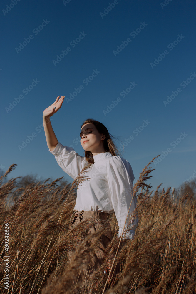 Young woman in white blouse standing in field of dry pampas grass in front of sky covering face by hand of bright sunlight. Style and fashion. Girl in casual outfit. Golden hour Young woman in white blouse standing in field of dry pampas grass in front of sky covering face by hand of bright sunlight. Style and fashion. Girl in casual outfit. Golden hour