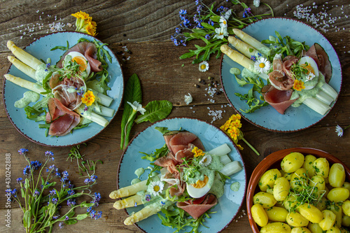 seasonal dinner with asparagus, prosciutto and young potatoes on a plate, Frankfurt green sauce, lunch with fresh wild herbs and flowers, eggs, wood dark background 