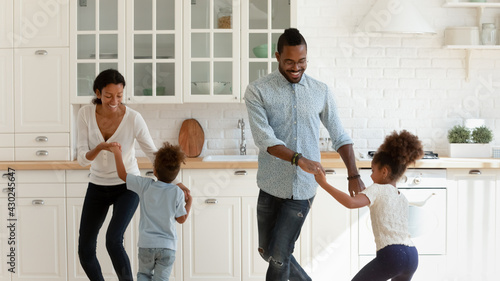 Happy African American parents and two kids dancing to music in kitchen, holding hands, having fun and laughing. Excited active parents and children exercising, enjoying family party in new house