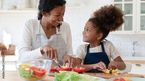 Happy mom and daughter kid talking and laughing while cooking dinner in kitchen together. Sweet preschooler girl helping mom to prepare salad from fresh vegetables  cutting organic pepper on table