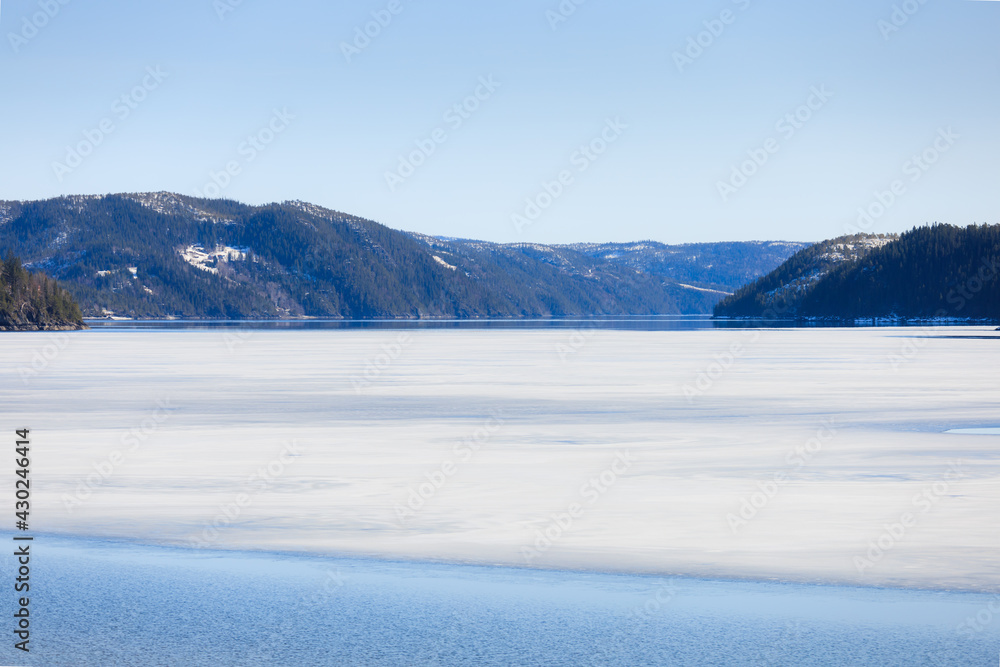 Lake thaw of Spring landscape. Forest lake partially covered with ice and partially reflected the blue sky and mountains, at the beginning of a spring day.