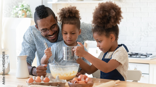 Happy dad teaching two cute preschooler kids to bake. Children and daddy beating eggs and sifting flour into bowl for kneading dough, making pie, cookies together. Family cooking, home bakery concept