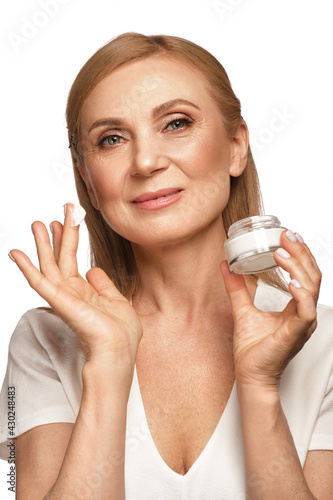 Portrait of a beautiful elderly woman in a white shirt with a moisturizing face cream in her hands.