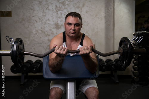 an adult man with a barbell shakes his biceps in the gym while sitting on a scott bench