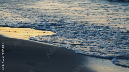 Front view of waves striking to the shore. Sand textured Beach in the foreground. The Shot is taken during the sunset. Orange reflection of the sun can be seen. Beautiful Calm Beach 