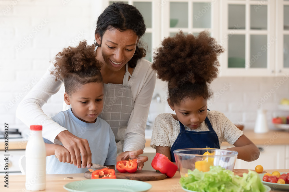 Happy mom teaching two preschooler kids to make vegetarian meal, slicing fresh pepper for salad. Mother and children cooking vegetables together in modern kitchen, keeping healthy eating diet