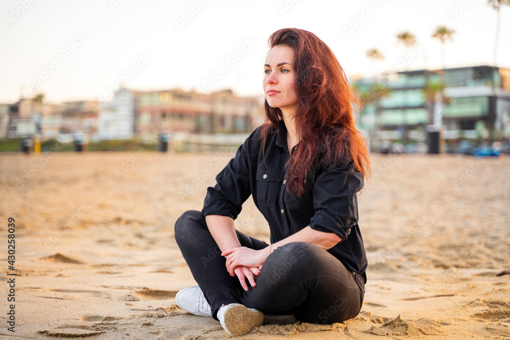 A modern young woman sits on the sand in Manhattan Beach in the early morning