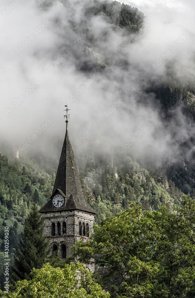 a church with a big clock in a mountain village in the Pyrenees with clouds behind it coming down the mountainside, church of Arties, Lleida, Spain, vertical