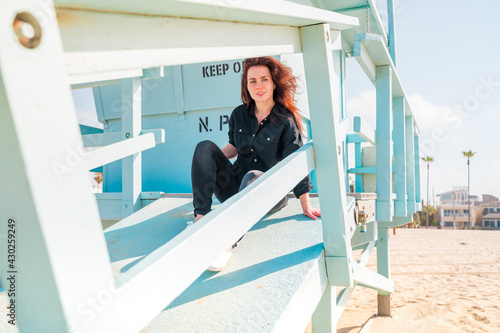 Beautiful young woman with long hair in a black shirt poses on a light blue lifeguard tower on a Los Angeles beach