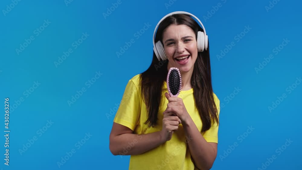 European woman singing and dancing with hair brush or comb instead microphone at blue studio background. Lady in headphones having fun, listening to music, dreams of being celebrity. European woman singing and dancing with hair brush or comb instead microphone at blue studio background. Lady in headphones having fun, listening to music, dreams of being celebrity.