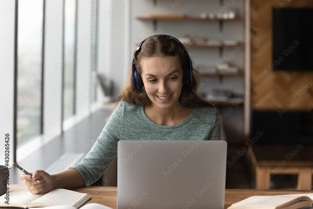 Happy young woman in headphones look at laptop screen make notes write in notebook consult client or customer online. Smiling female study distant on computer, talk on webcam video zoom call. Happy young woman in headphones look at laptop screen make notes write in notebook consult client or customer online. Smiling female study distant on computer, talk on webcam video zoom call.