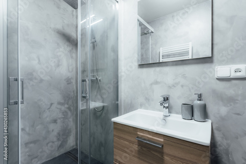 Contemporary bathroom with shower and sink with wooden cabinet under it and mirror with integrated light. All walls are painted with concrete trowel in gray. Whole space gives modern impression.