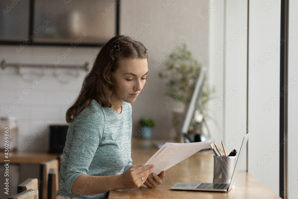 Focused millennial Caucasian female employee work online on laptop consider paper document or contract. Young woman worker busy using computer read paper correspondence in office workplace. Focused millennial Caucasian female employee work online on laptop consider paper document or contract. Young woman worker busy using computer read paper correspondence in office workplace.