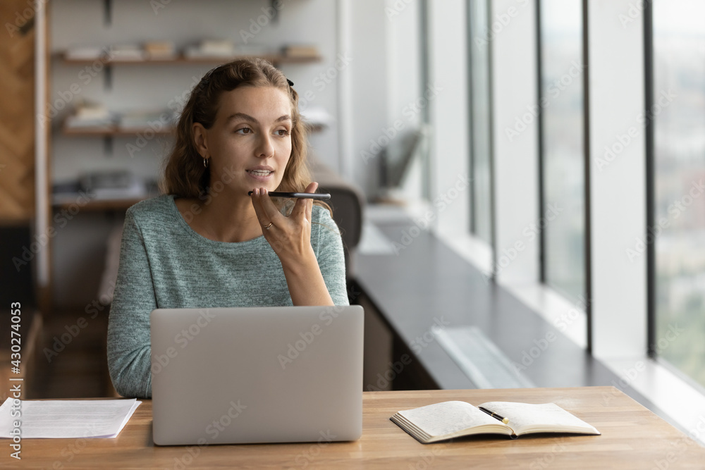 Young Caucasian businesswoman sit at desk in office work online on computer record audio voice message on smartphone. Female employee consult client on cellphone, talk speak on loudspeaker on gadget. Young Caucasian businesswoman sit at desk in office work online on computer record audio voice message on smartphone. Female employee consult client on cellphone, talk speak on loudspeaker on gadget.