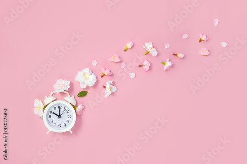 Alarm clock and flying spring flowers and petals on pink background. Springtime concept. Flat lay, place for text.