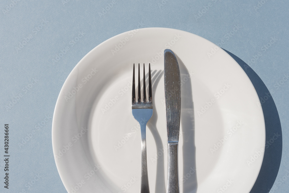Summer outdoor eating concept - cropped closeup of white plate and knife and fork on blue background with hard shadows (selective focus)