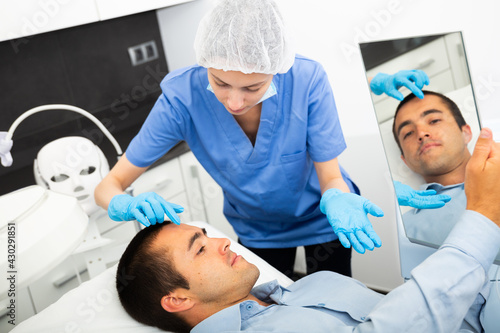 Skilled woman beautician consulting male client before facial treatment in medical esthetic office.