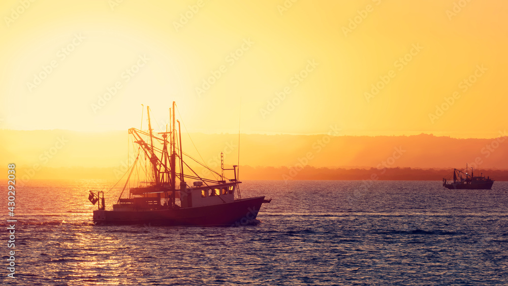 Silhouette of a Trawler Fishing Boat heading out to sea. Dramatic sunset with beautiful golden light. Commercial Fishing Industry concept. 