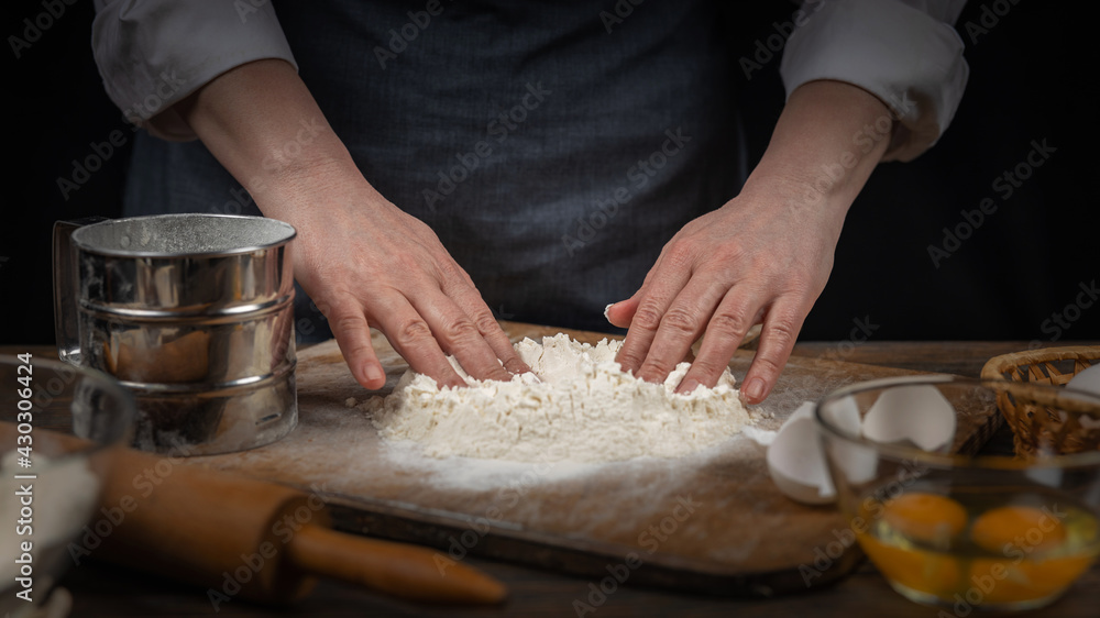 Women's hands, flour and dough. A woman in an apron prepares dough for homemade baking, a rustic home cozy atmosphere, a dark background with unusual lighting.