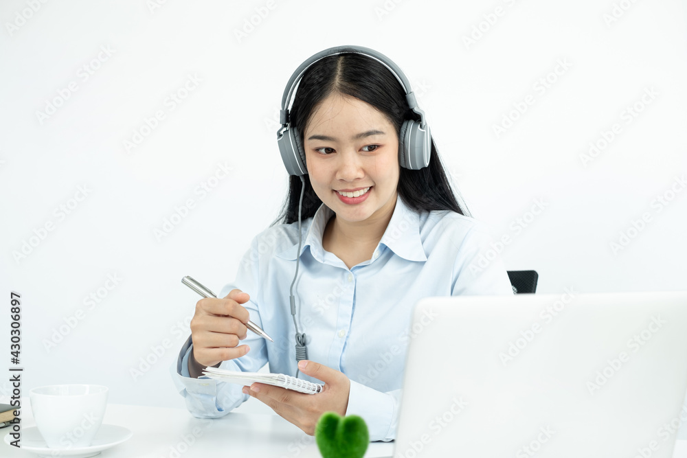 Video call, a young Asian woman wearing headphones, communicates via online video long distance, looking at the digital laptop screen, happily greeted with a friend Video call, a young Asian woman wearing headphones, communicates via online video long distance, looking at the digital laptop screen, happily greeted with a friend