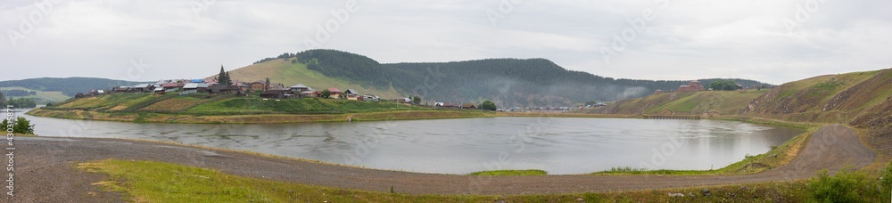 Small cozy village stands on shore of lake or river, hills and fog in background. Panoramic view. Early cool morning, overcast gray sky