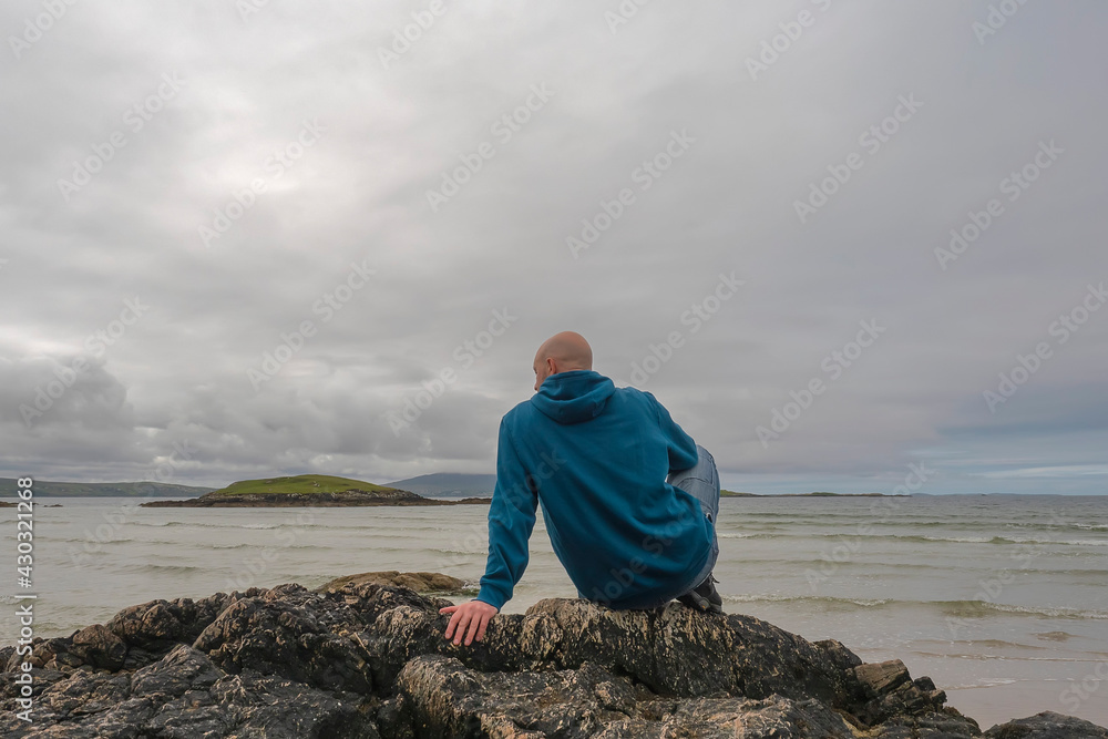 Bald man sitting on a rock by the ocean looking water. Beautiful nature scenery in the background. West coast of Ireland. Back to camera. Cloudy sky. Bald man sitting on a rock by the ocean looking water. Beautiful nature scenery in the background. West coast of Ireland. Back to camera. Cloudy sky.