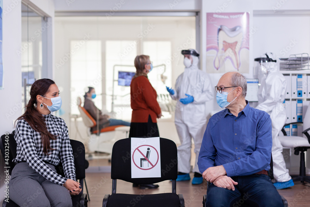 Obraz premium Senior man with face mask discussing with woman patient in stomatology clinic in waiting room, keeping social distancing during global pandemic with coronavirus. Doctor standing wearing ppe suit.