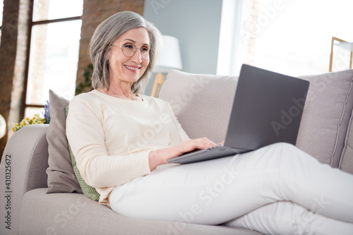 Photo of cheerful happy nice old woman lie sofa use computer wear glasses smile indoors inside house home