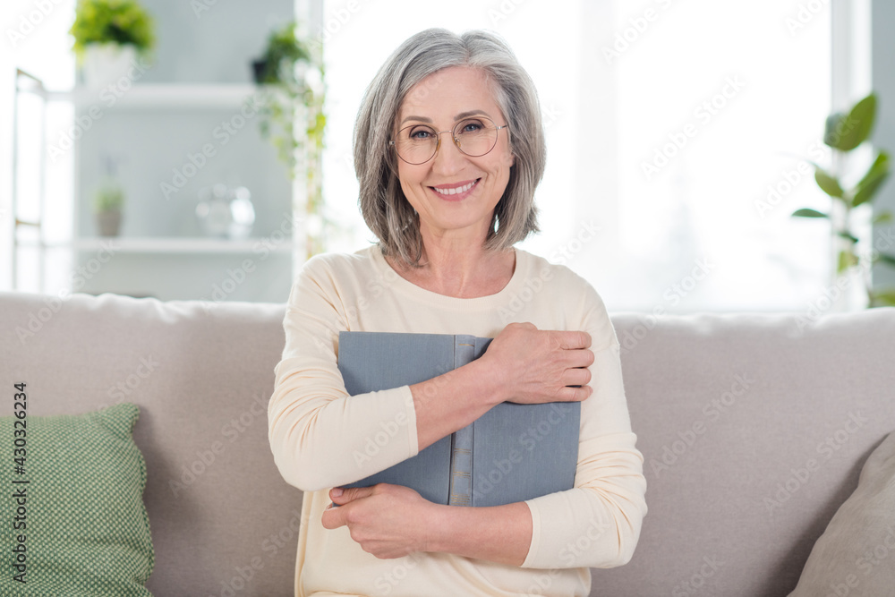 Photo of charming positive happy old lady hug book literature smile indoors inside house home apartment Photo of charming positive happy old lady hug book literature smile indoors inside house home apartment