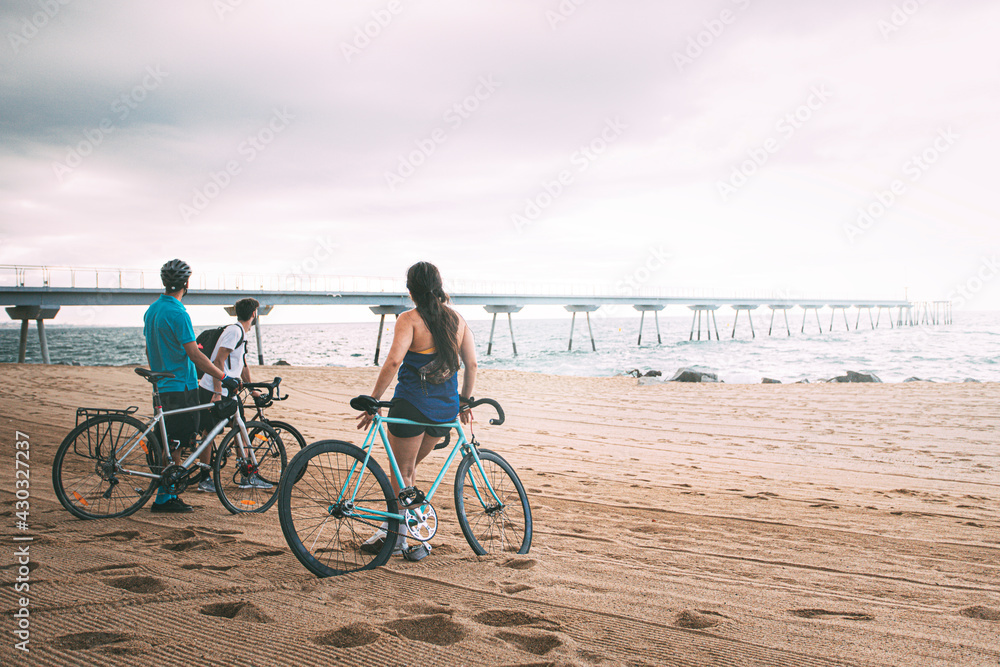 Obraz premium Friends seen from behind with their bikes on the beach at dawn looking out to sea. Young people enjoying their free time outdoors. background with copyspace. Concept of free time. Concept of sport