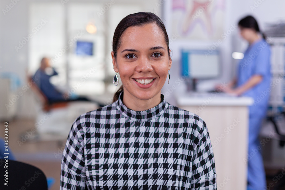 Obraz premium Portrait of smiling woman patient looking on camera sitting on chair in waiting room of stomatological clinic. Stomatologist assistant typing on pc in dental office while doctor working in background.