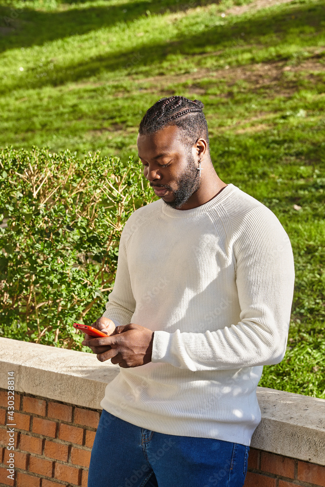 Fototapeta premium Handsome african american guy chatting via smartphone standing, cheerful dark-skinned man enjoying free time blogging in networks via smartphone.