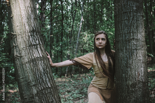 Confident, beautiful woman in a native american dress with face paint standing in the woods between trees, nature concept, protect the forest concept