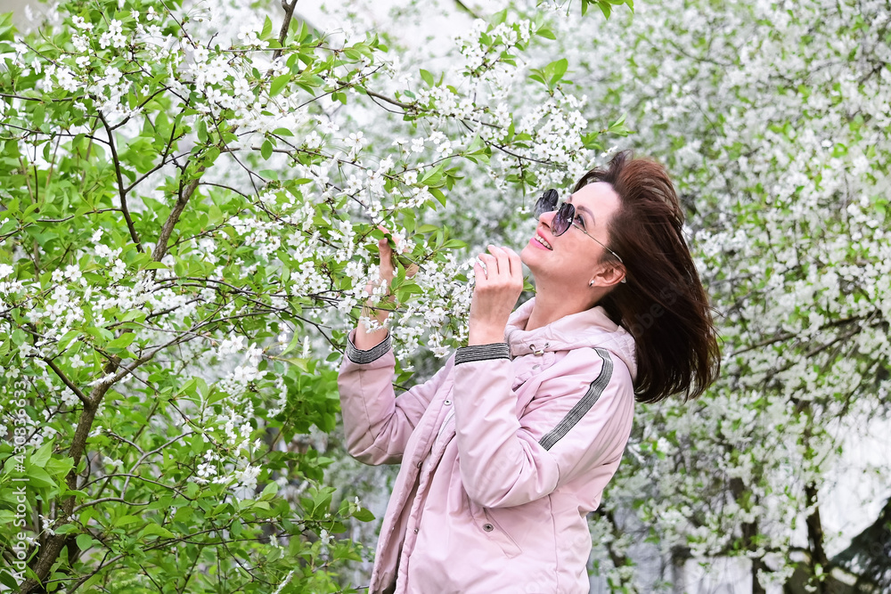 A stylish woman in sunglasses poses among the blooming spring cherry branches in the open air, a calm moment. Portrait of a fashionable brunette in a pink jacket in white colors A stylish woman in sunglasses poses among the blooming spring cherry branches in the open air, a calm moment. Portrait of a fashionable brunette in a pink jacket in white colors
