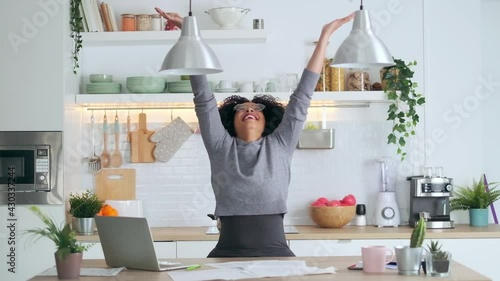 Video of happy afro business woman tossing invoices and documents into the air while working on the laptop at home.