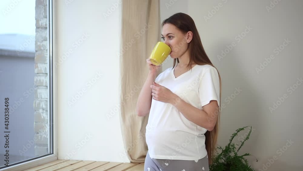 Pregnant woman drinking coffee and standing by window in apartment room spbd. Pregnant woman drinking coffee and standing by window in apartment room spbd.