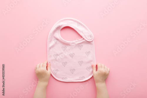 Baby girl hands holding textile bib with heart shapes for feeding on light pink table background. Pastel color. Closeup. Point of view shot. Top down view.