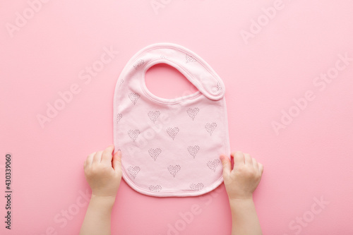 Baby girl hands holding textile bib with heart shapes for feeding on light pink table background. Pastel color. Closeup. Point of view shot. Top down view. photo