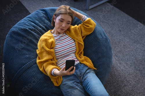 Asian businesswoman chilling in office relaxing space lying in beanbag looking at smartphone