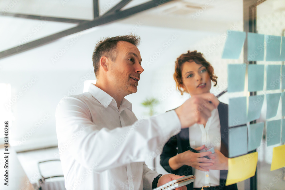 Obraz premium Discussing ideas. Two business people talking while looking at post it notes on a glass wall.