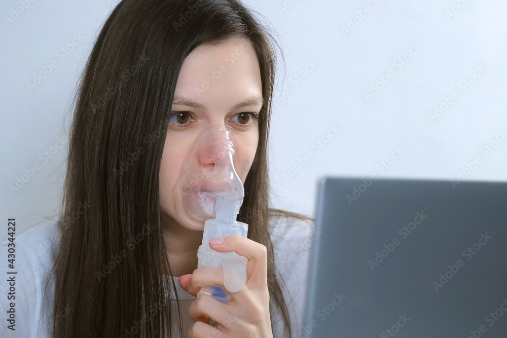 Sick young woman is using nebulizer and inhaler for treatment at home and working on laptop. She is breathing in mask. Fibrosis, covid-19, asthma, copd. Treatment, cure and procedure, therapy. Sick young woman is using nebulizer and inhaler for treatment at home and working on laptop. She is breathing in mask. Fibrosis, covid-19, asthma, copd. Treatment, cure and procedure, therapy.