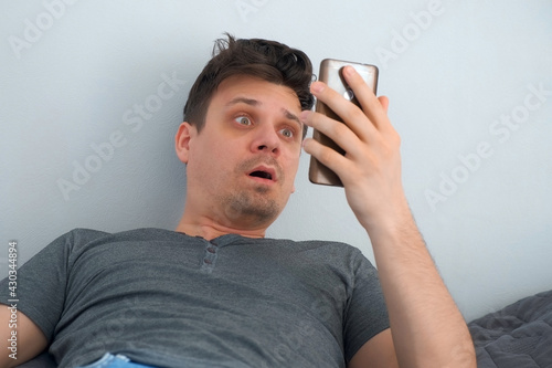Man with hangover waking up in his bed, looking at time on smartphone and very surprised. He yawns and clutches his head which acking. Headache, drug and alcoholism addiction, human bad habits.
