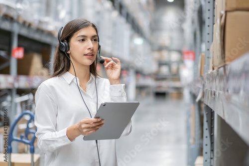 Portrait of warehouse woman staff using digital tablet and talking to customer with headset working in distribution factory. female call center or support operator working for online shopping.