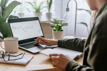 Distance learning online education and work. Business woman with plants writes in notebook. Cropped freelance girl with laptop at home office. Using computer with economics graphics and diagrams.