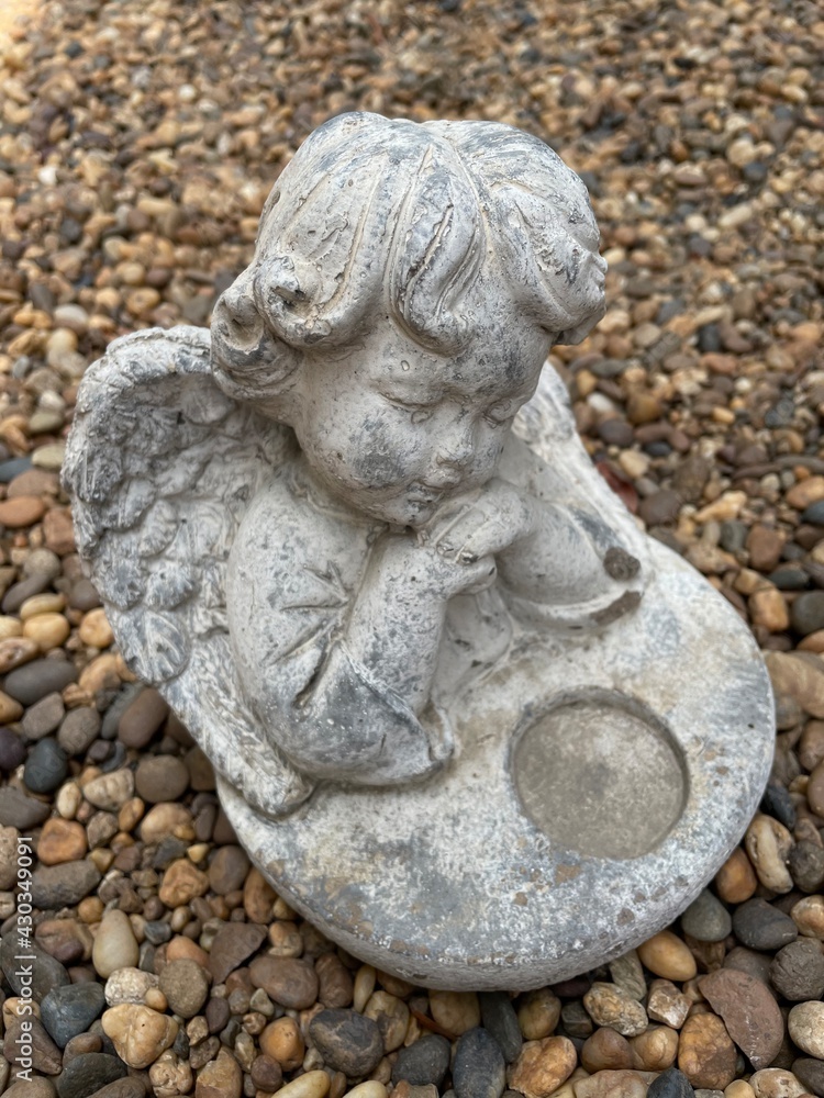 Little cute cupid angel sculpture on river gravel background in the english garden. Little cute cupid angel sculpture on river gravel background in the english garden.