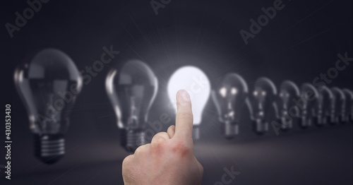 Composition of finger touching lit light bulb with row of light bulbs on black background