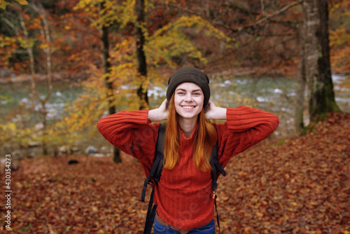 joyful woman in a hat sweater with a backpack on her back gestures with her hands in a park in nature