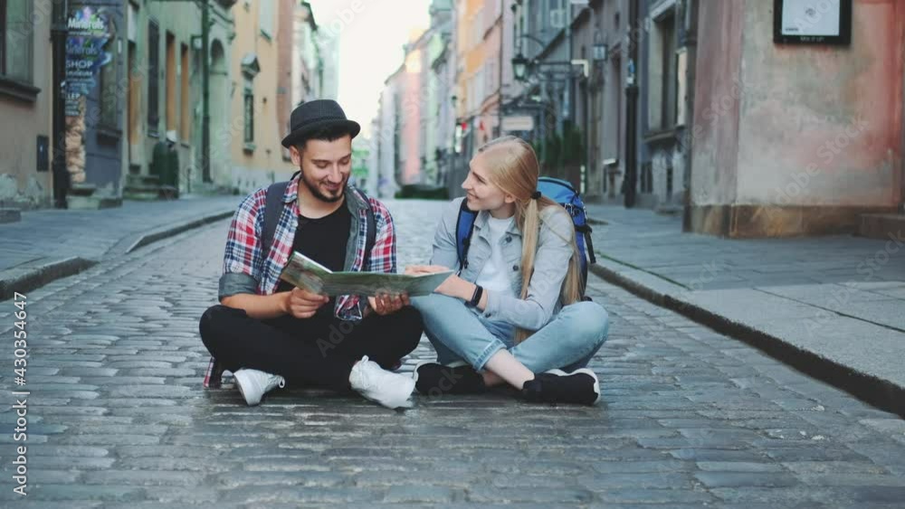 Young tourists couple using map, sitting on pavement and admiring historical surroundings Young tourists couple using map, sitting on pavement and admiring historical surroundings