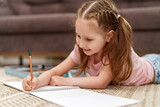 Little cute Caucasian girl child, drawing in an album with colored pencils, lying on floor. hardworking child does homework. The child's hobby is drawing. new modern set for drawing and creativity